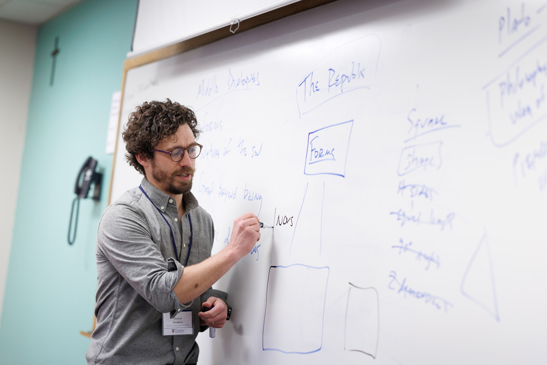 Man with glasses writing on a whiteboard in a classroom, surrounded by notes and diagrams. He appears focused and engaged in teaching.