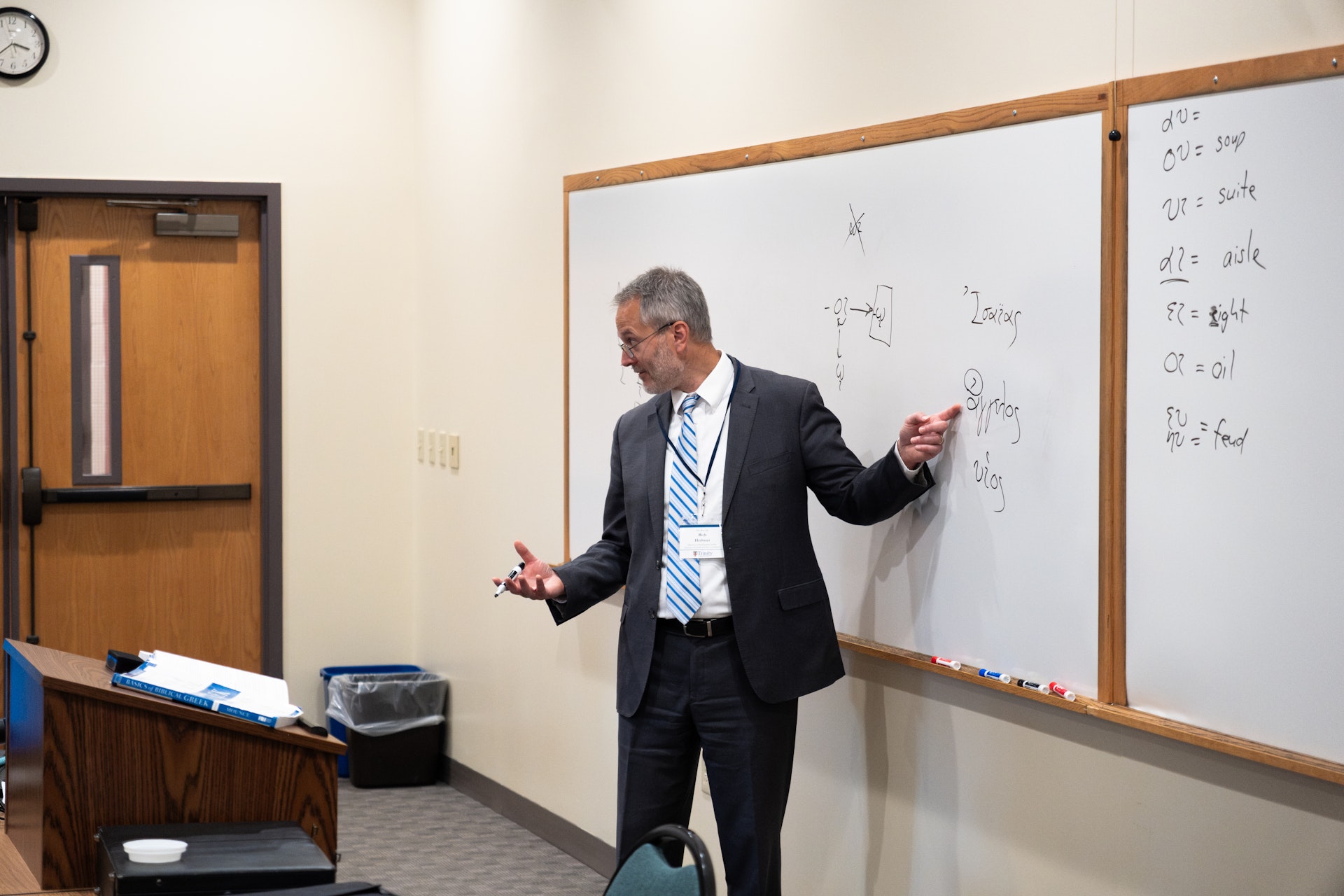 A man in a suit gestures while giving a presentation in a classroom. He stands beside a whiteboard with diagrams and written notes. The room is minimal, featuring a clock, a door, and a podium. The atmosphere conveys a professional and educational tone.