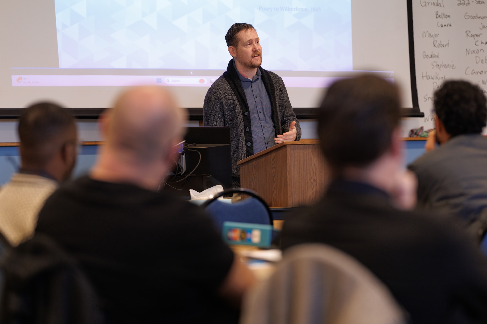 A man is speaking at a podium in a classroom, with a projection screen behind him. Attentive audience members are seated, facing the speaker.