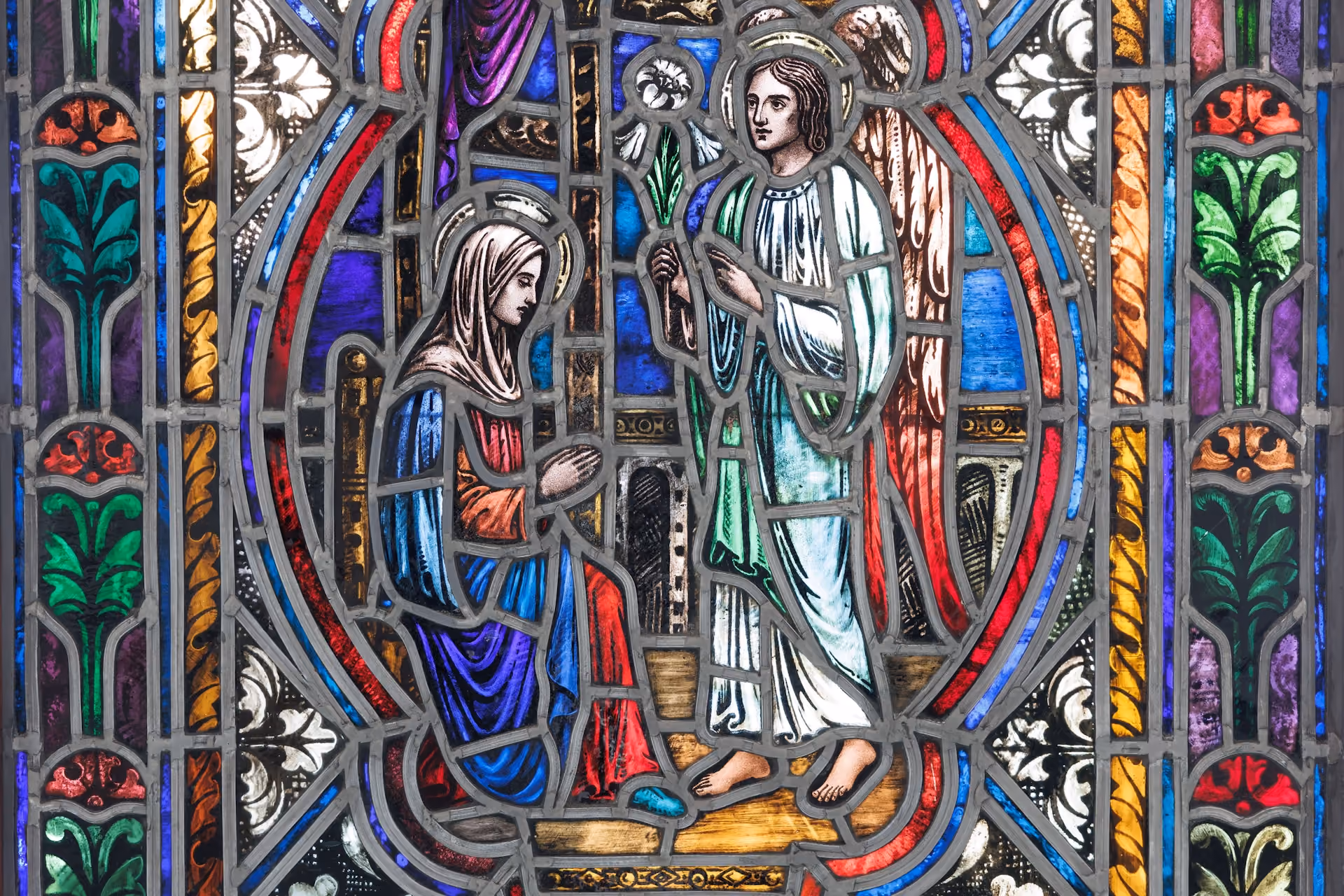 Stained glass depicting a kneeling woman in blue and red robes facing an angel in white with wings, holding a lily, framed by vibrant colors.