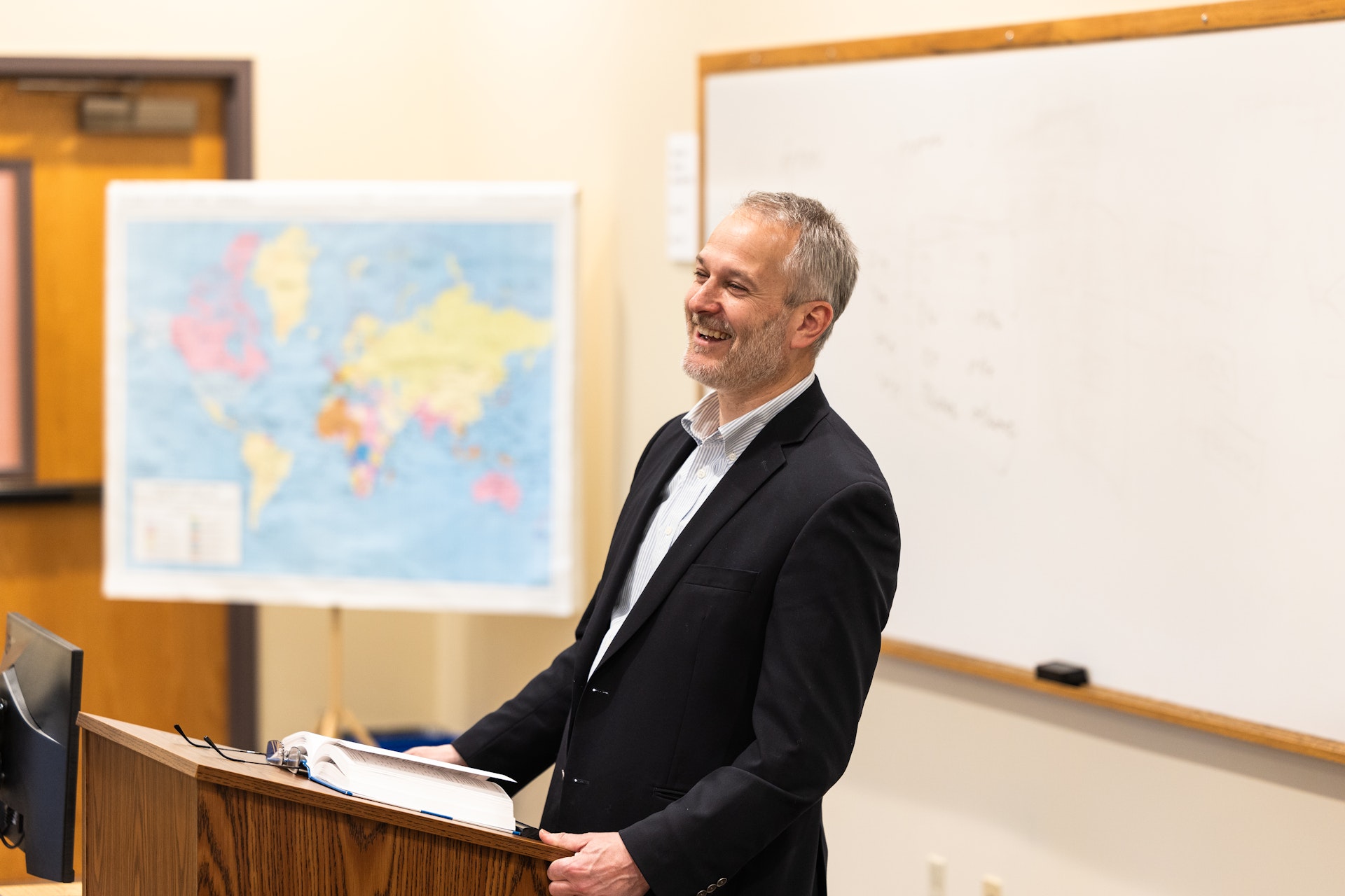 A smiling man in a suit stands at a lectern in a classroom, speaking. A map on an easel and a whiteboard are visible in the background.