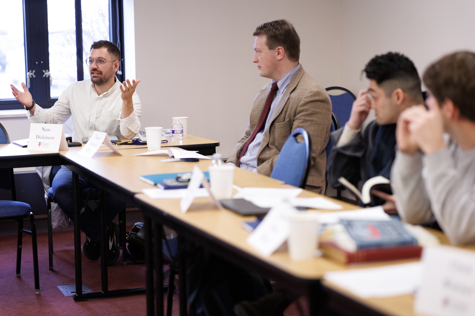 A group of people sit around a U-shaped table in a meeting room. One man gestures while speaking, as others attentively listen and take notes. The atmosphere is focused and collaborative.