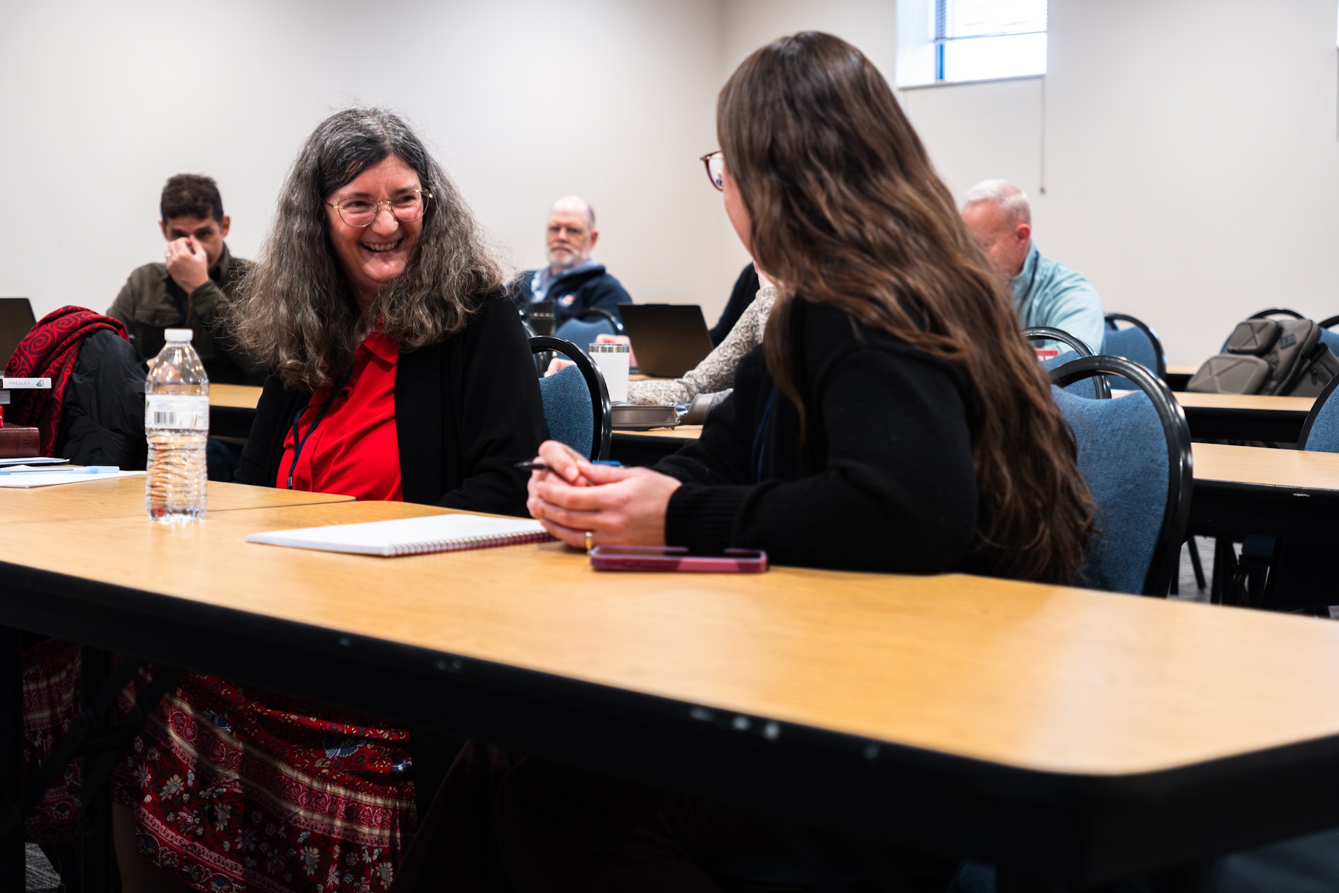 Two women engaging in a lively conversation at a classroom table. The setting is informal, with other people working on laptops in the background.