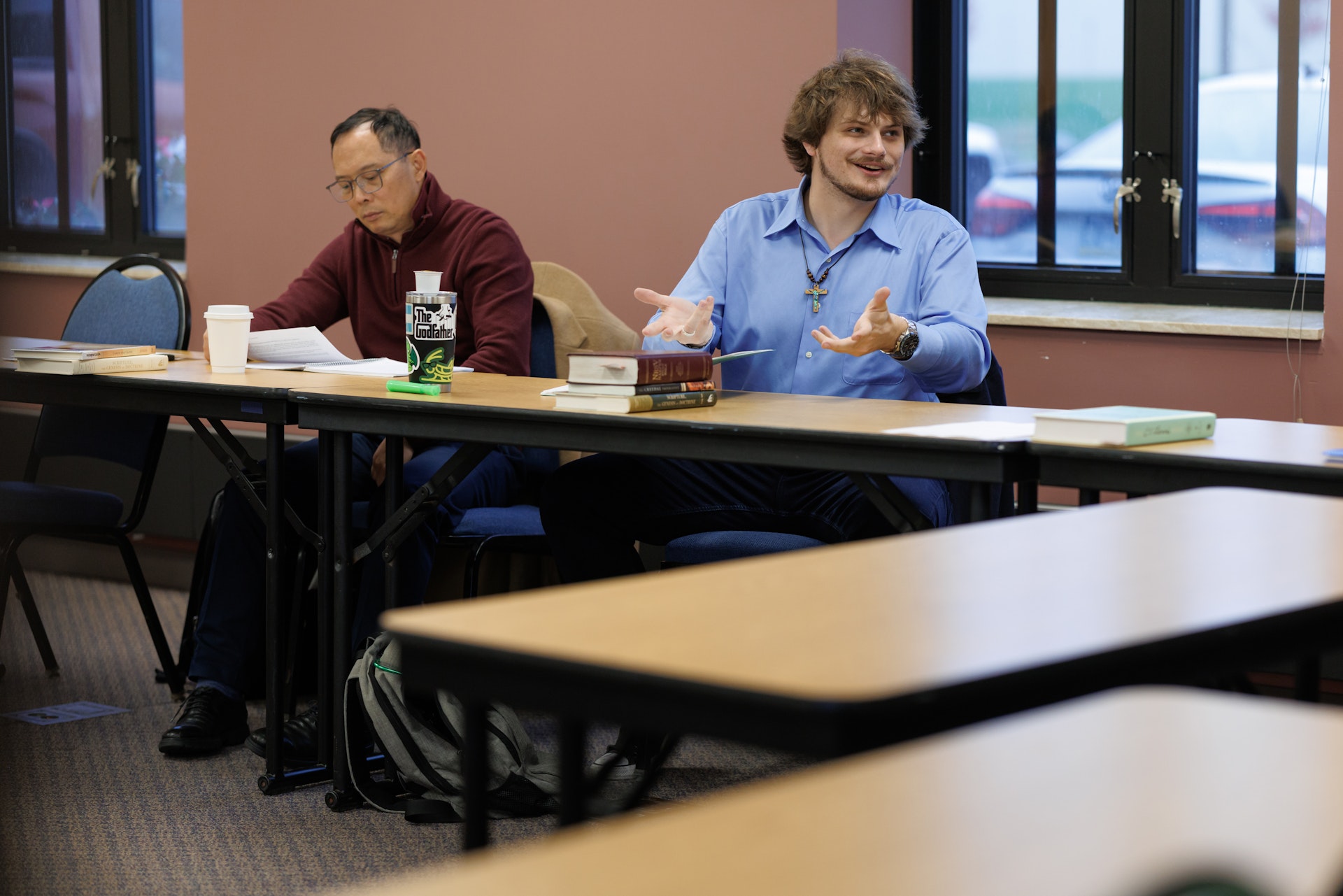 Two people are seated at a long table in a classroom. One man in a blue shirt is speaking energetically, while the other is focused on reading.