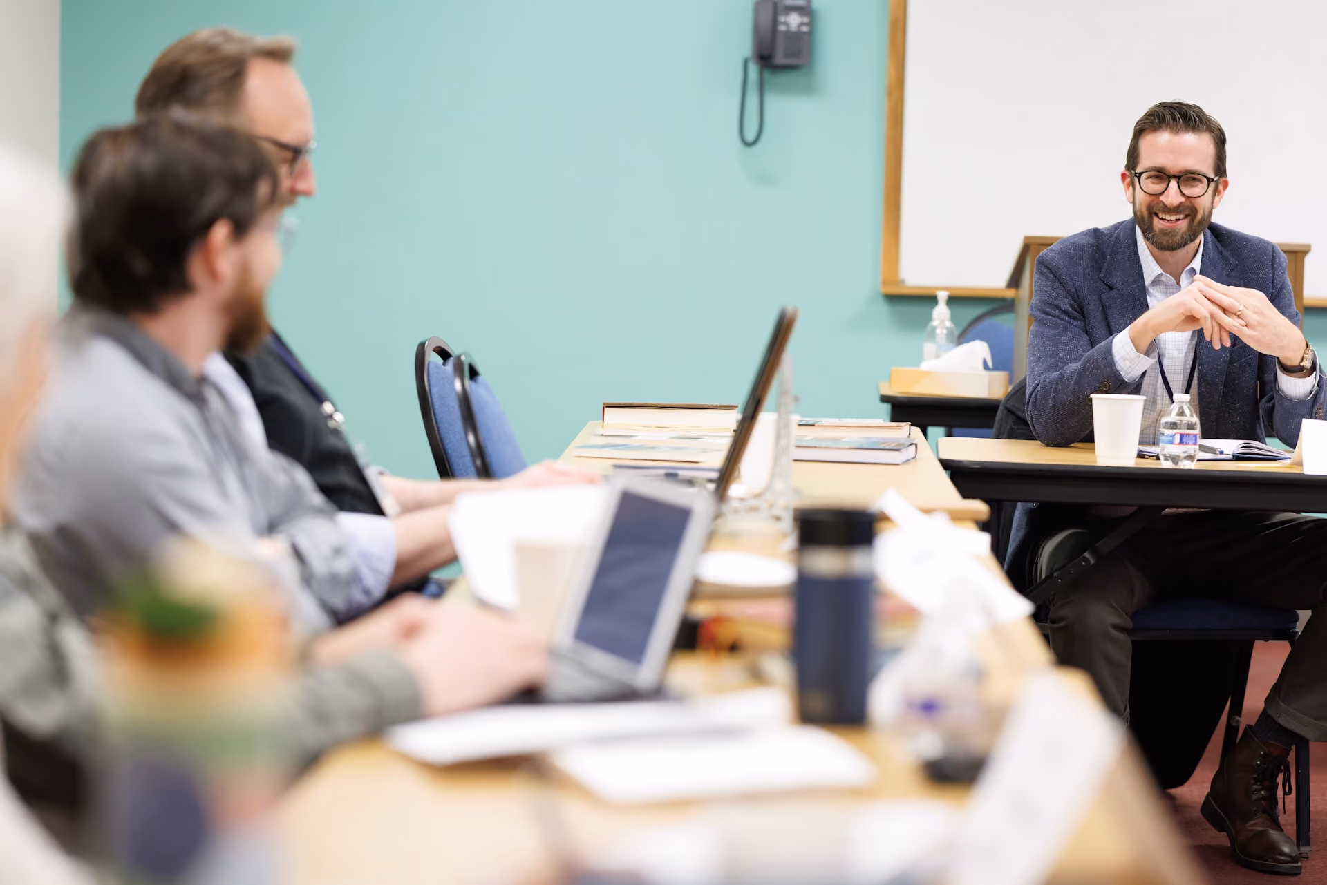 A group of people in a meeting room, with a man in a suit smiling and speaking. Others listen attentively with laptops open, creating a collaborative atmosphere.