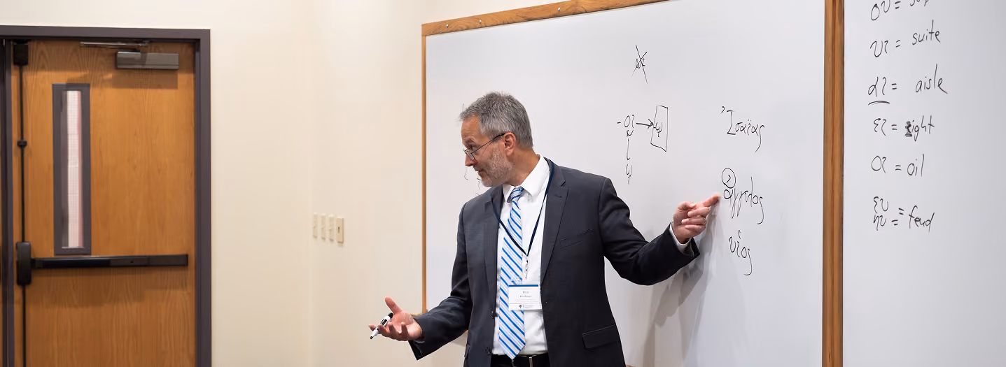 A man in a suit gestures while giving a presentation in a classroom. He stands beside a whiteboard with diagrams and written notes. The room is minimal, featuring a clock, a door, and a podium. The atmosphere conveys a professional and educational tone.