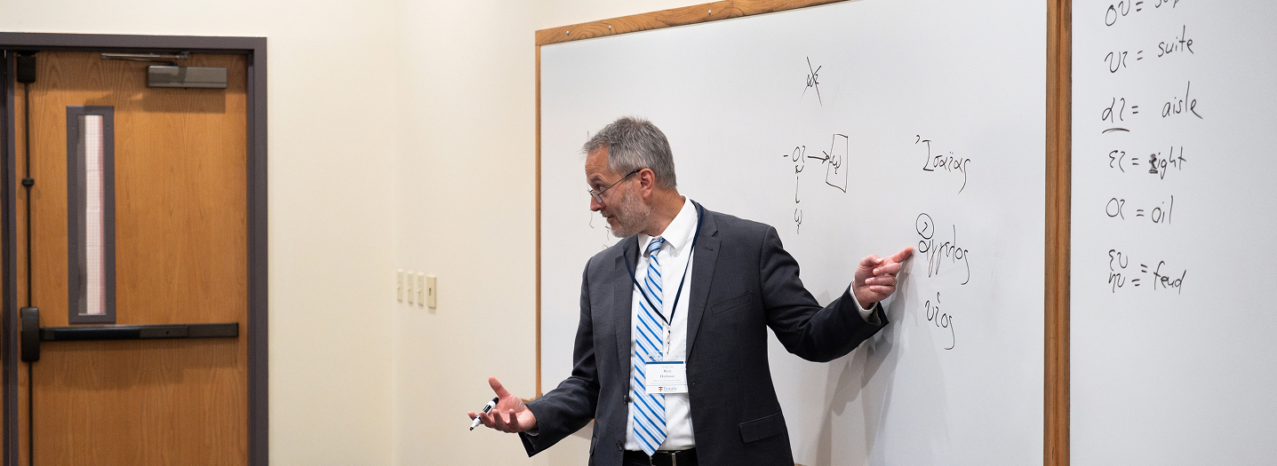 A man in a suit gestures while giving a presentation in a classroom. He stands beside a whiteboard with diagrams and written notes. The room is minimal, featuring a clock, a door, and a podium. The atmosphere conveys a professional and educational tone.