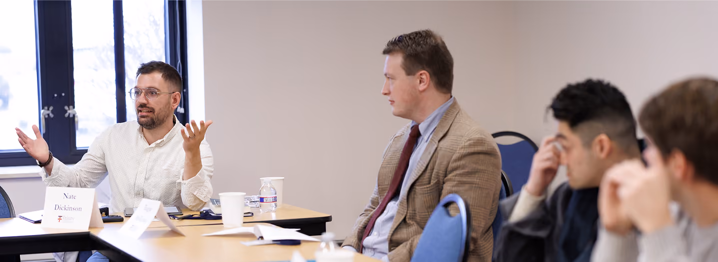 A group of men engaged in a discussion around a conference table. One man gestures while talking, with others listening intently. Papers and coffee cups are on the table.