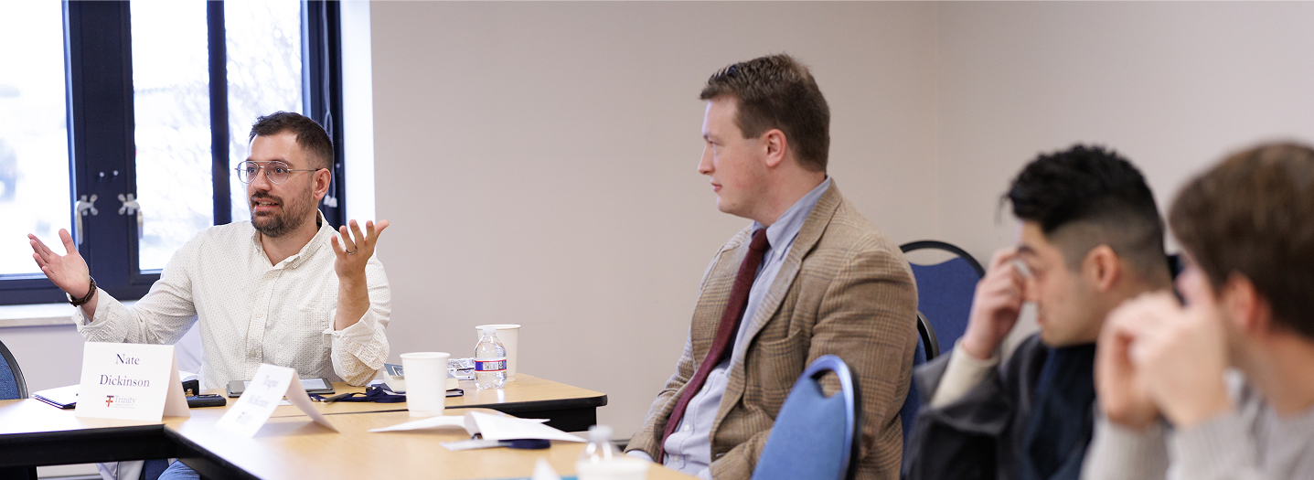 A group of men engaged in a discussion around a conference table. One man gestures while talking, with others listening intently. Papers and coffee cups are on the table.