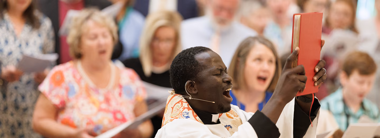 A joyful priest in white vestments, holding up a red book, sings passionately with a diverse choir in the background during a lively church service.