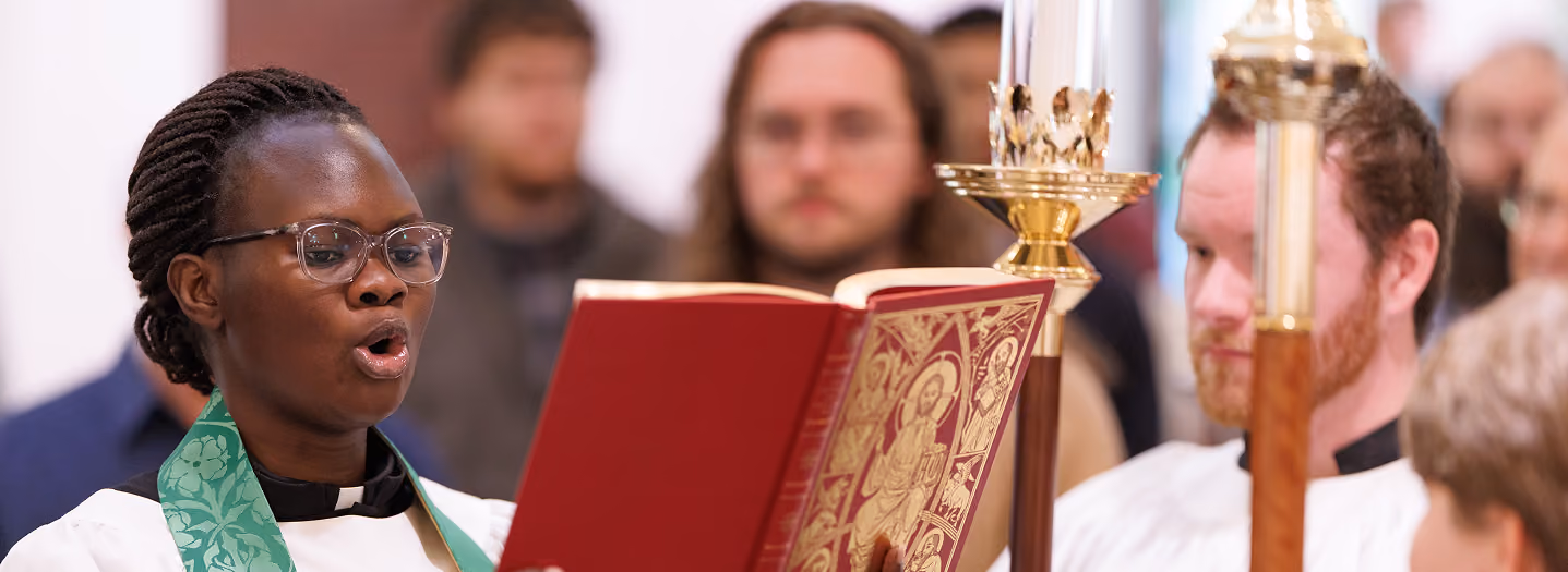 A person in religious robes sings from an ornate red book, surrounded by others in white vestments, conveying a solemn and reverent atmosphere.