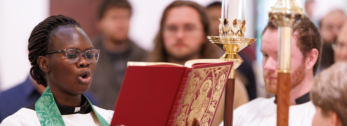 A person in religious robes sings from an ornate red book, surrounded by others in white vestments, conveying a solemn and reverent atmosphere.