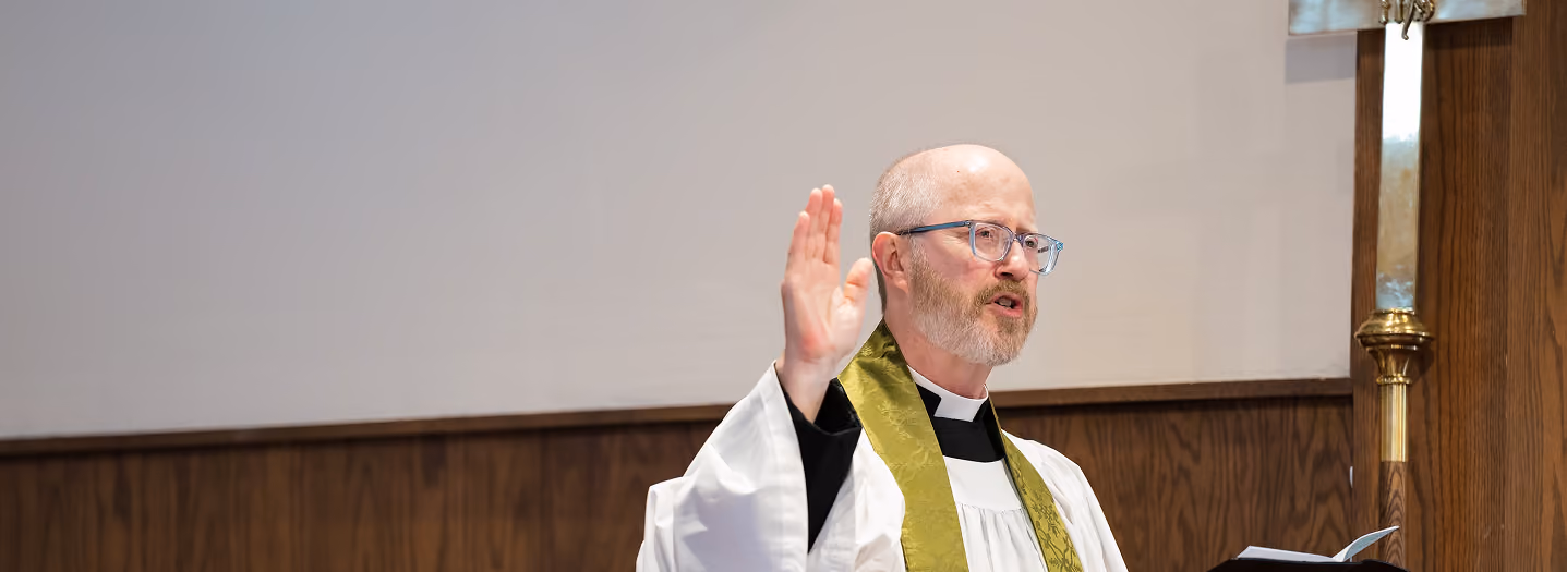 A priest in white robes with a green stole gestures while holding an open book, standing by a wooden cross in a church setting, conveying a solemn tone.