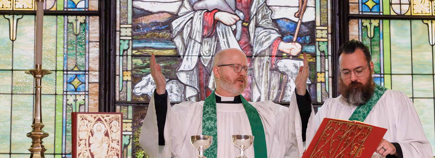Two clergy members in green stoles lead a church service at the altar, with a stained glass window of Jesus holding a lamb in the background. The atmosphere is solemn and reverent.