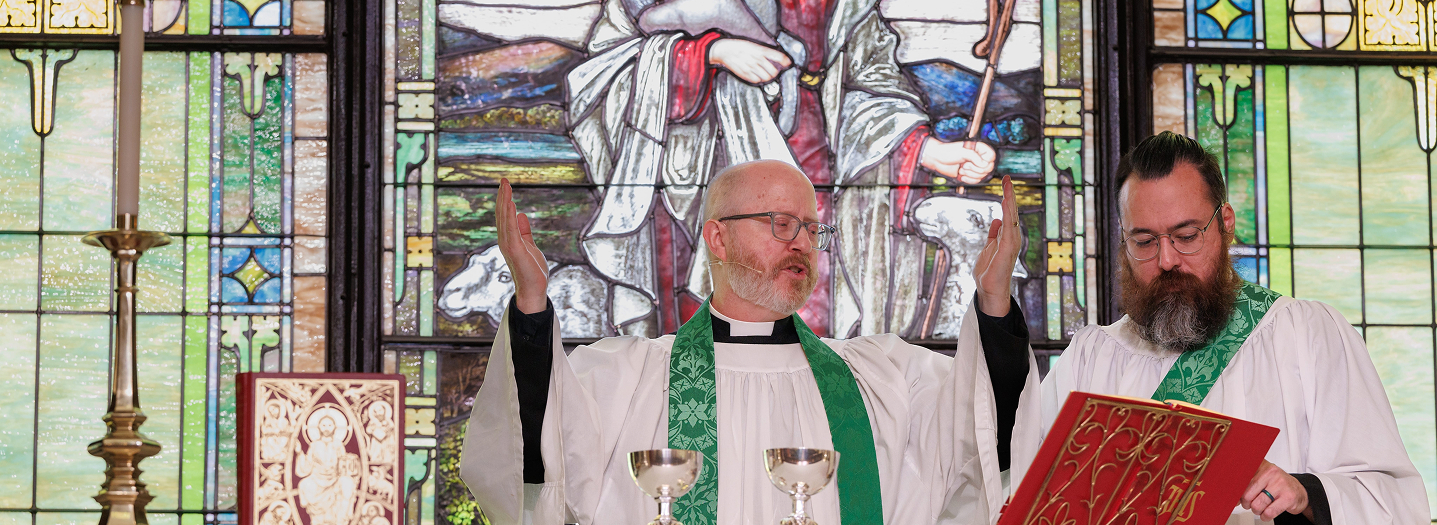 Two clergy members in green stoles lead a church service at the altar, with a stained glass window of Jesus holding a lamb in the background. The atmosphere is solemn and reverent.