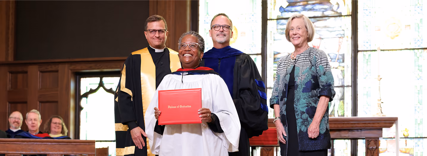 A woman in graduation attire is being hooded by a man in academic robes. They stand in a church-like setting, conveying a sense of achievement.