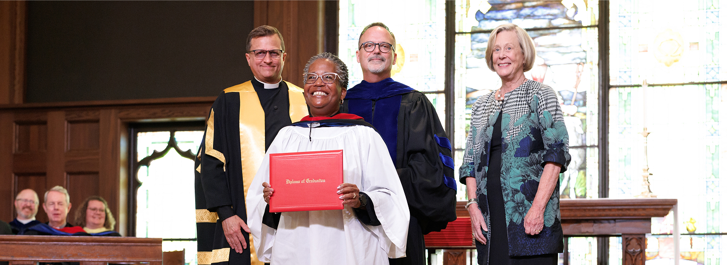 A woman in graduation attire is being hooded by a man in academic robes. They stand in a church-like setting, conveying a sense of achievement.