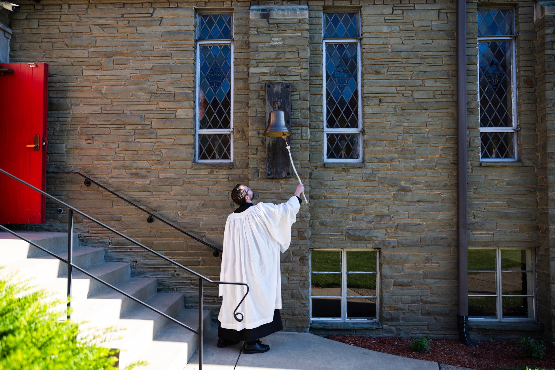 A person wearing a white surplice over a black cassock pulls a rope to ring a brass bell mounted on the stone wall of a chapel, beside leaded-glass windows and a red door.