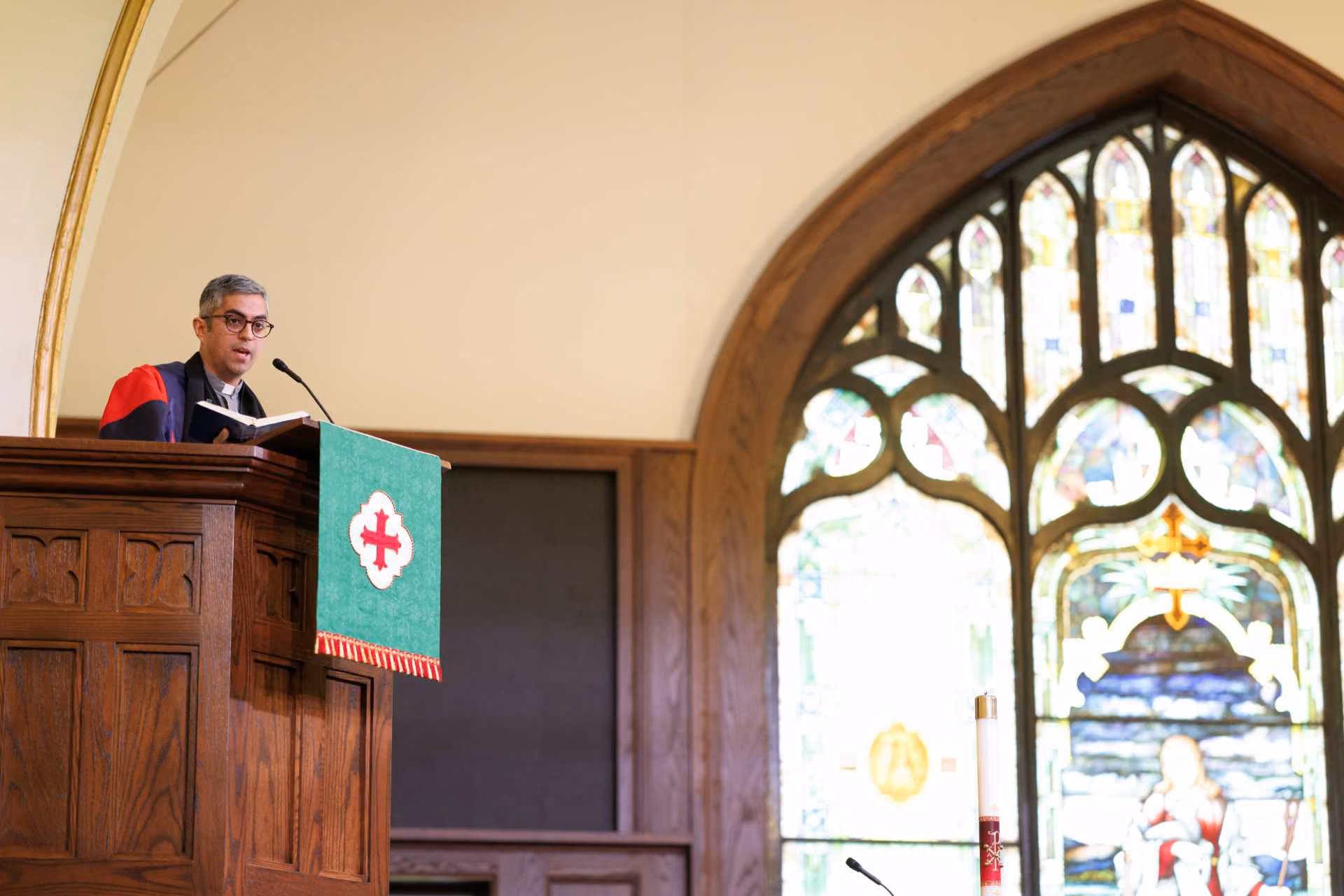 Seminary faculty member preaching at a wood pulpit in an Anglican chapel with a large stained glass window.