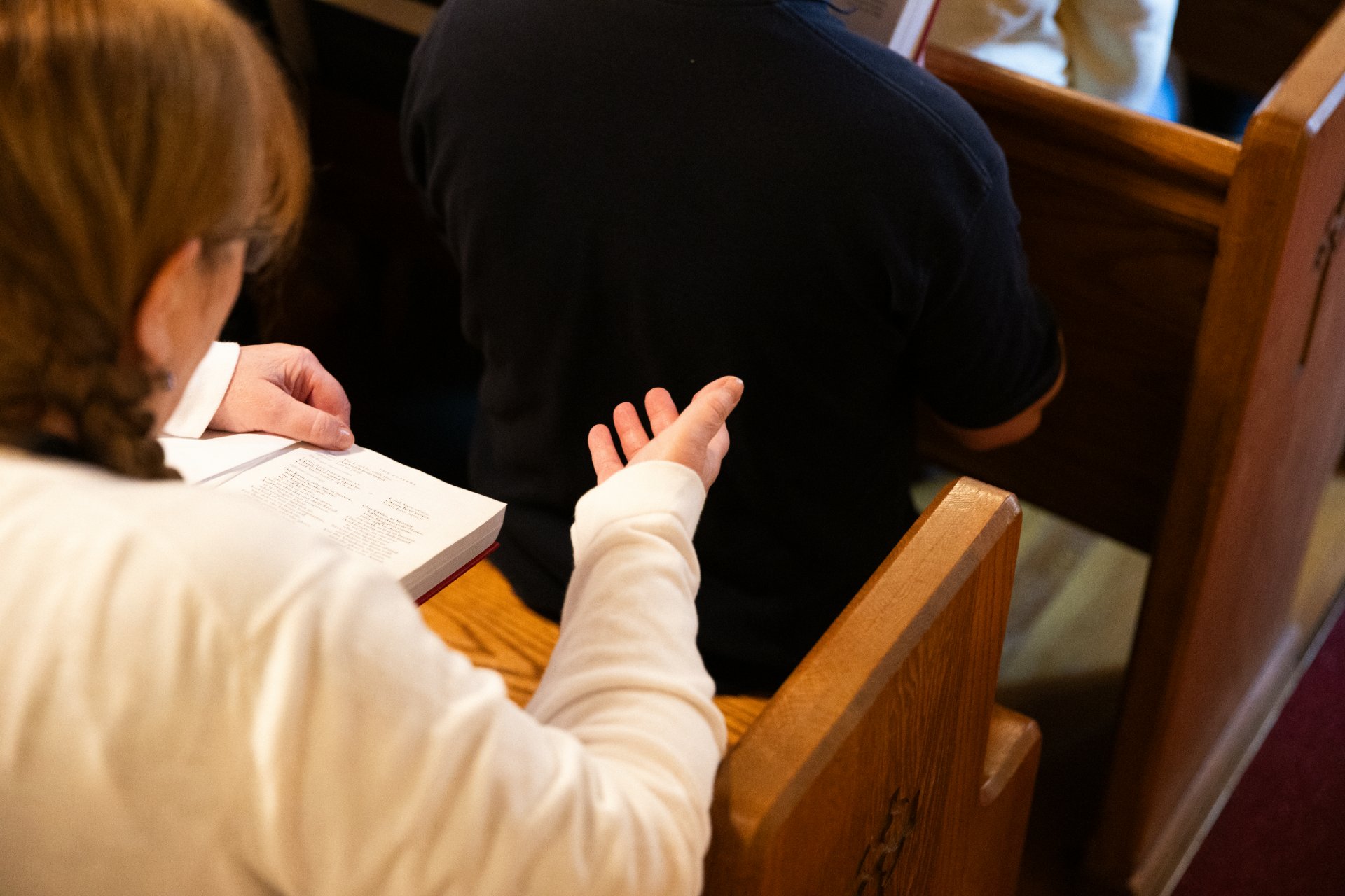 Viewed from above, a student in a white top holds an open liturgical book with red binding on the pew rail, hand raised in prayer