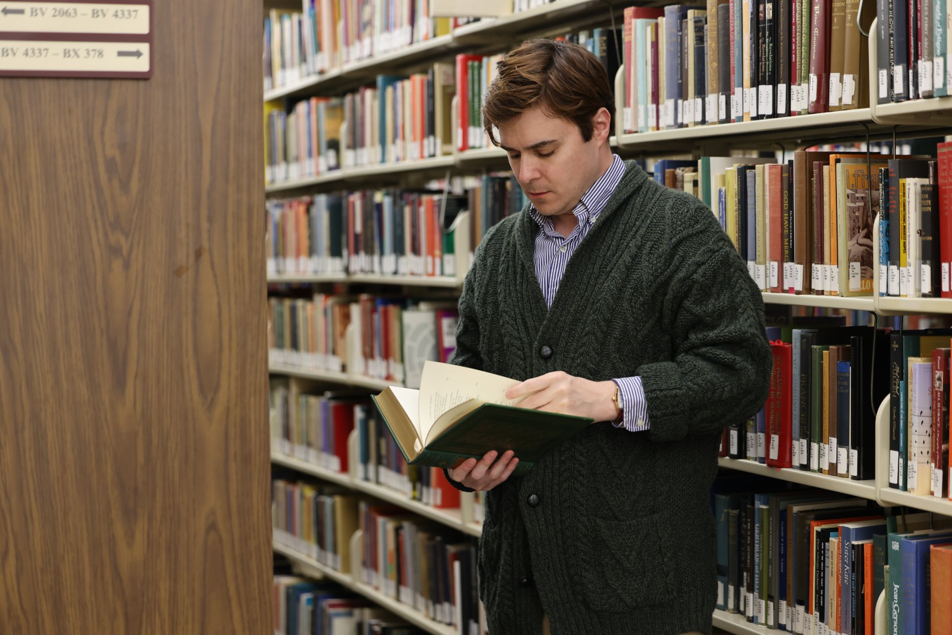 A young man in a cable-knit cardigan stands between two full bookshelves, reading an open book. Shelf signs show BV and BX call number ranges, indicating a theological library collection.
