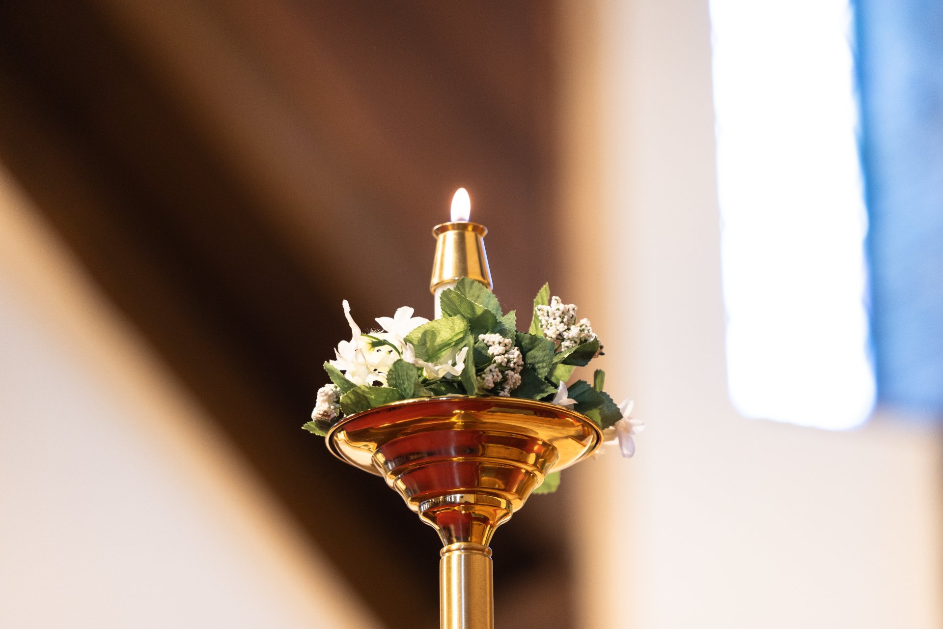 A close-up of a polished brass candlestick with a single burning candle, encircled by white floral decorations and green leaves. A stained glass window is softly visible in the blurred background.