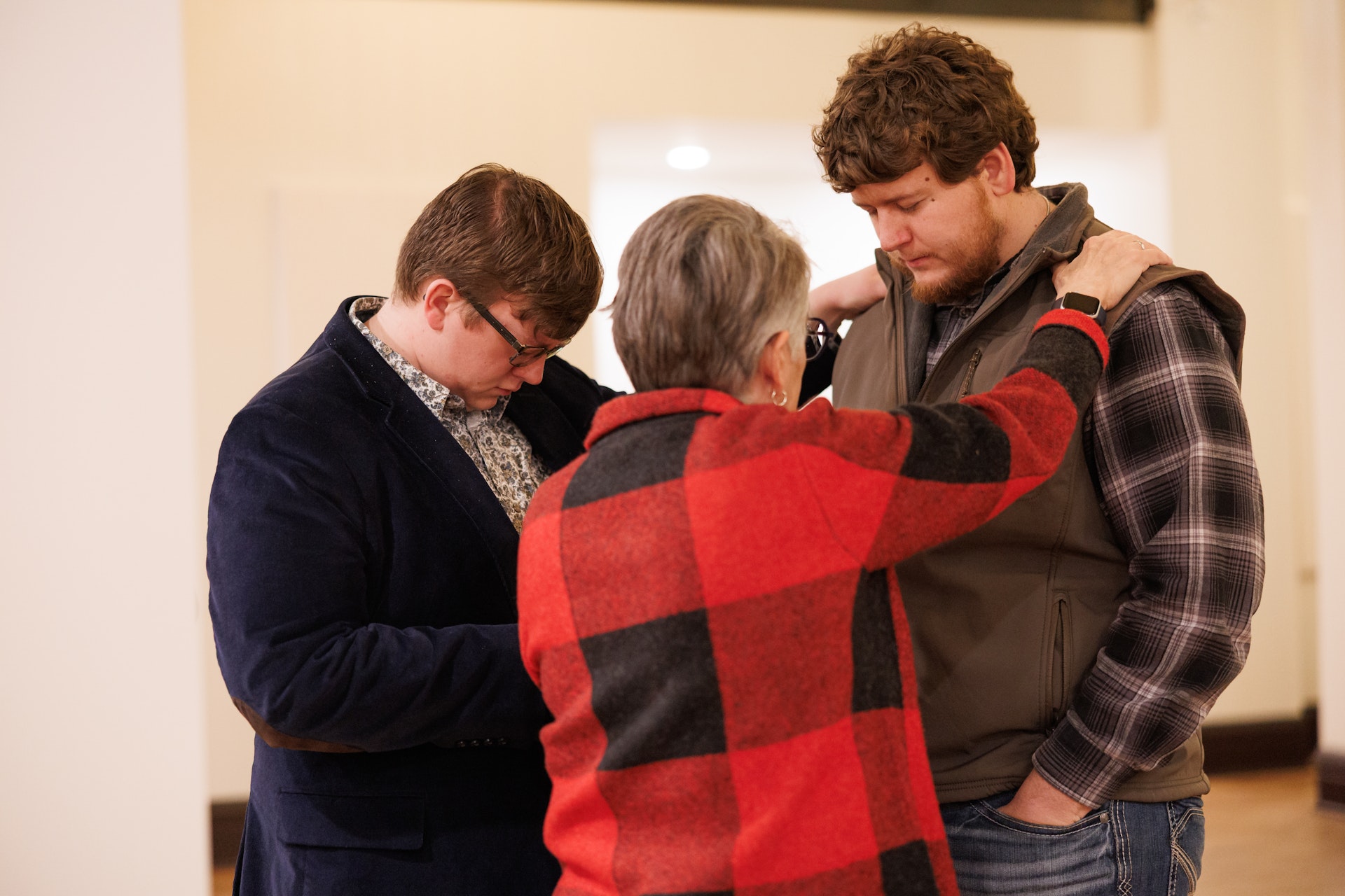 Three people stand together in an indoor setting, appearing to share an emotional or intimate moment. The person in a red plaid jacket extends arms to comfort the others.