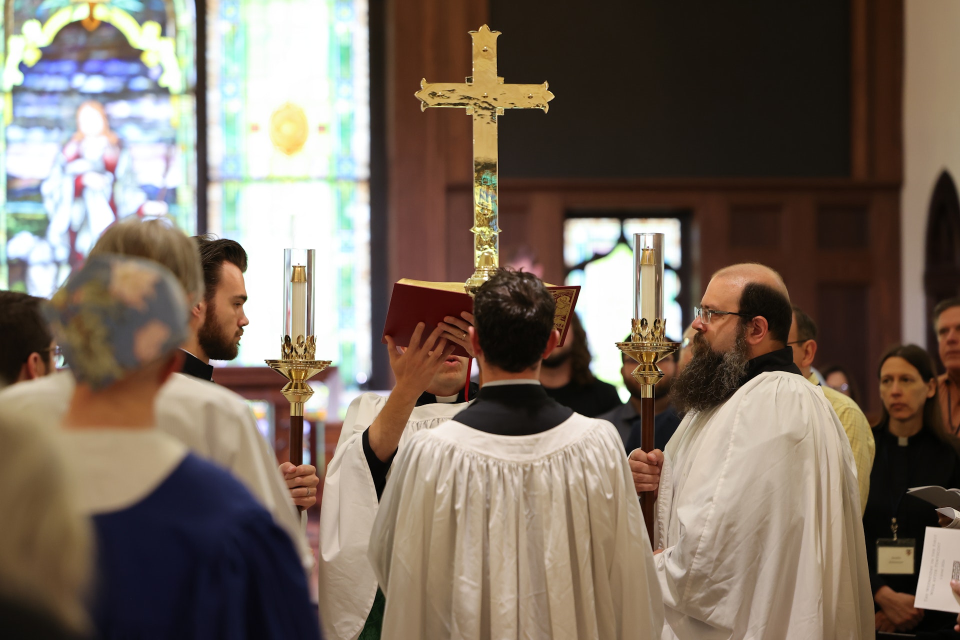 Robed acolytes bearing processional candles flank a minister elevating a red Gospel book, with a large brass processional cross leading the way through a wood-paneled church nave filled with congregants.