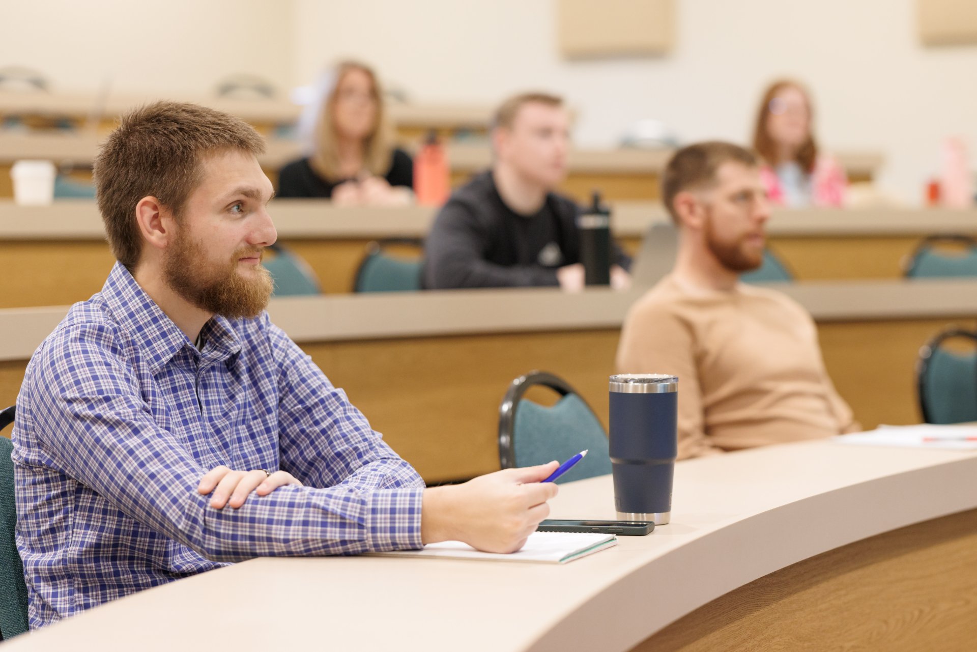 Several students sit at tiered lecture hall desks, notebooks and travel mugs on the desk surface, their attention directed toward an unseen instructor at the front of the room.