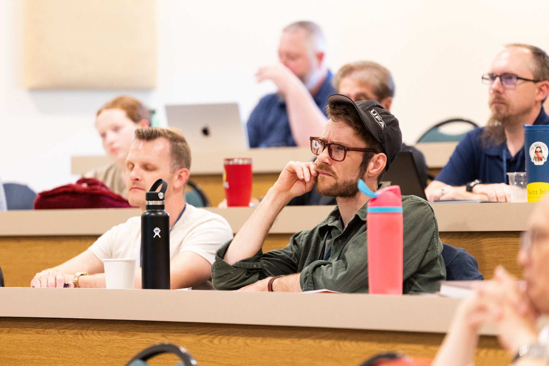Several adult students sit in tiered classroom rows, including a man in a cap and glasses in a focused, contemplative pose. Others look on or take notes, with personal items like a Hydro Flask and travel mugs visible on the desktops.