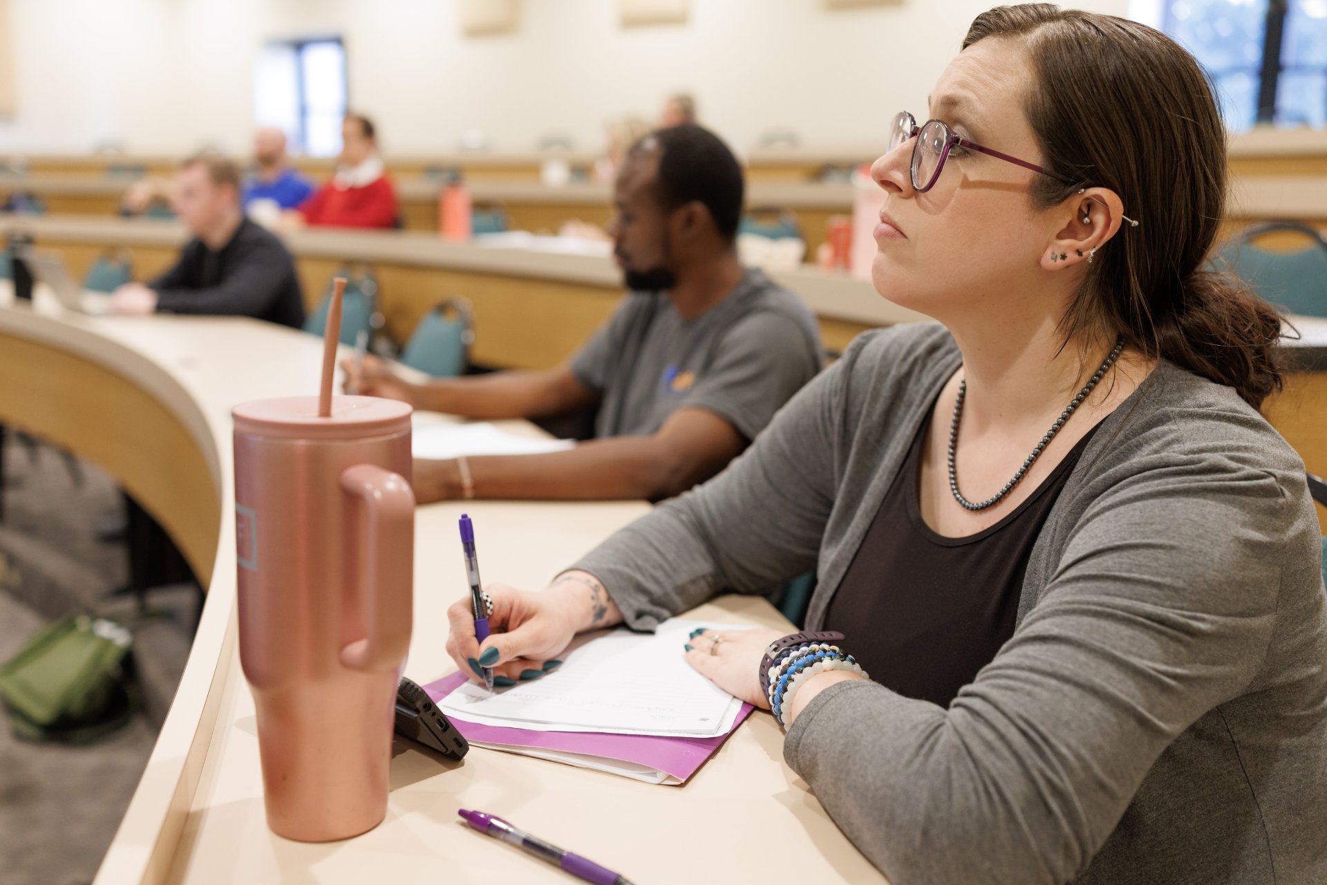 Seminary student takes notes during a theological education lecture, with classmates seated at tiered classroom desks behind her.