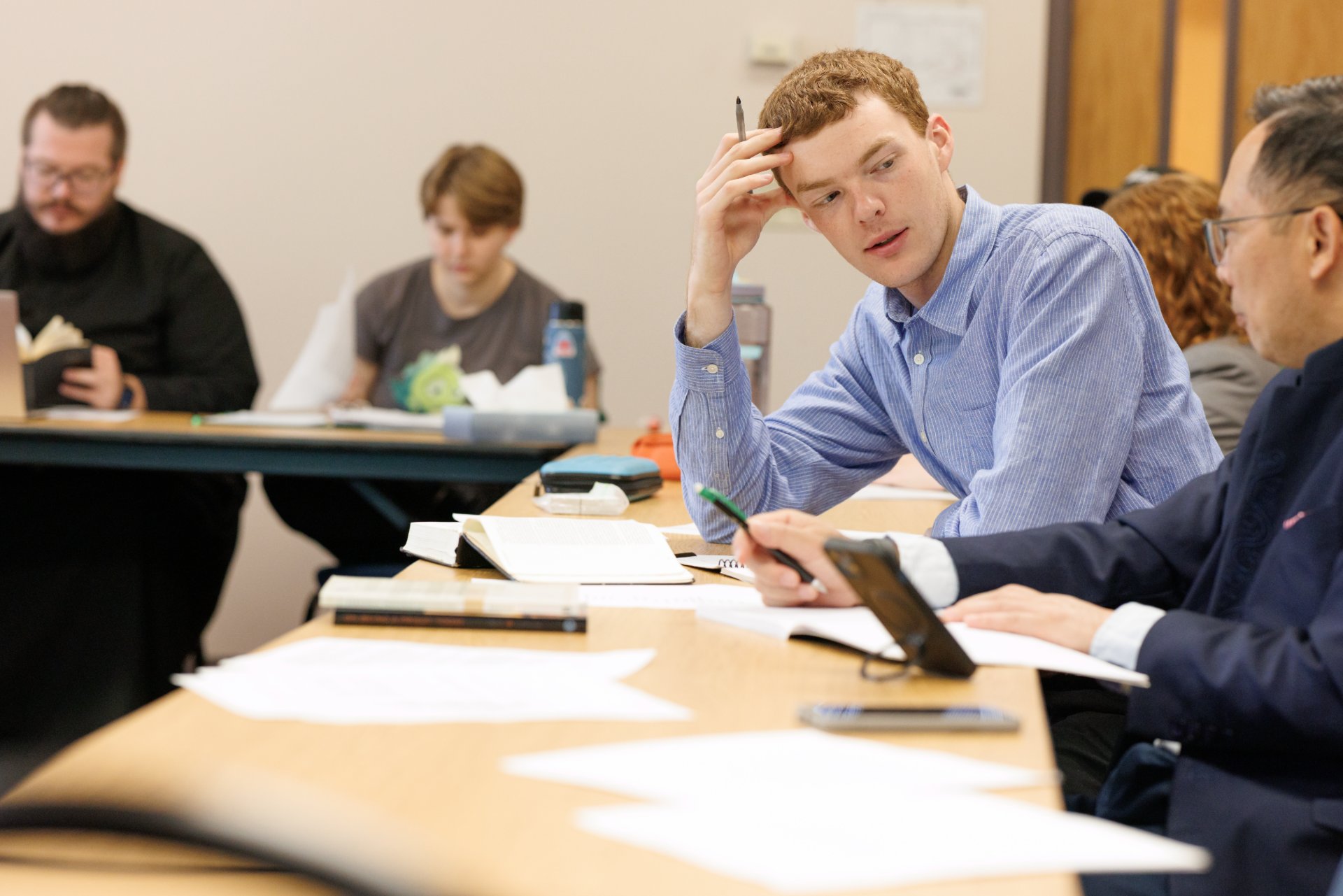 A young man in a blue shirt holds a pen to his forehead in focused thought while speaking with a fellow student at a shared table. In the background, two other students study independently with open books and notebooks.