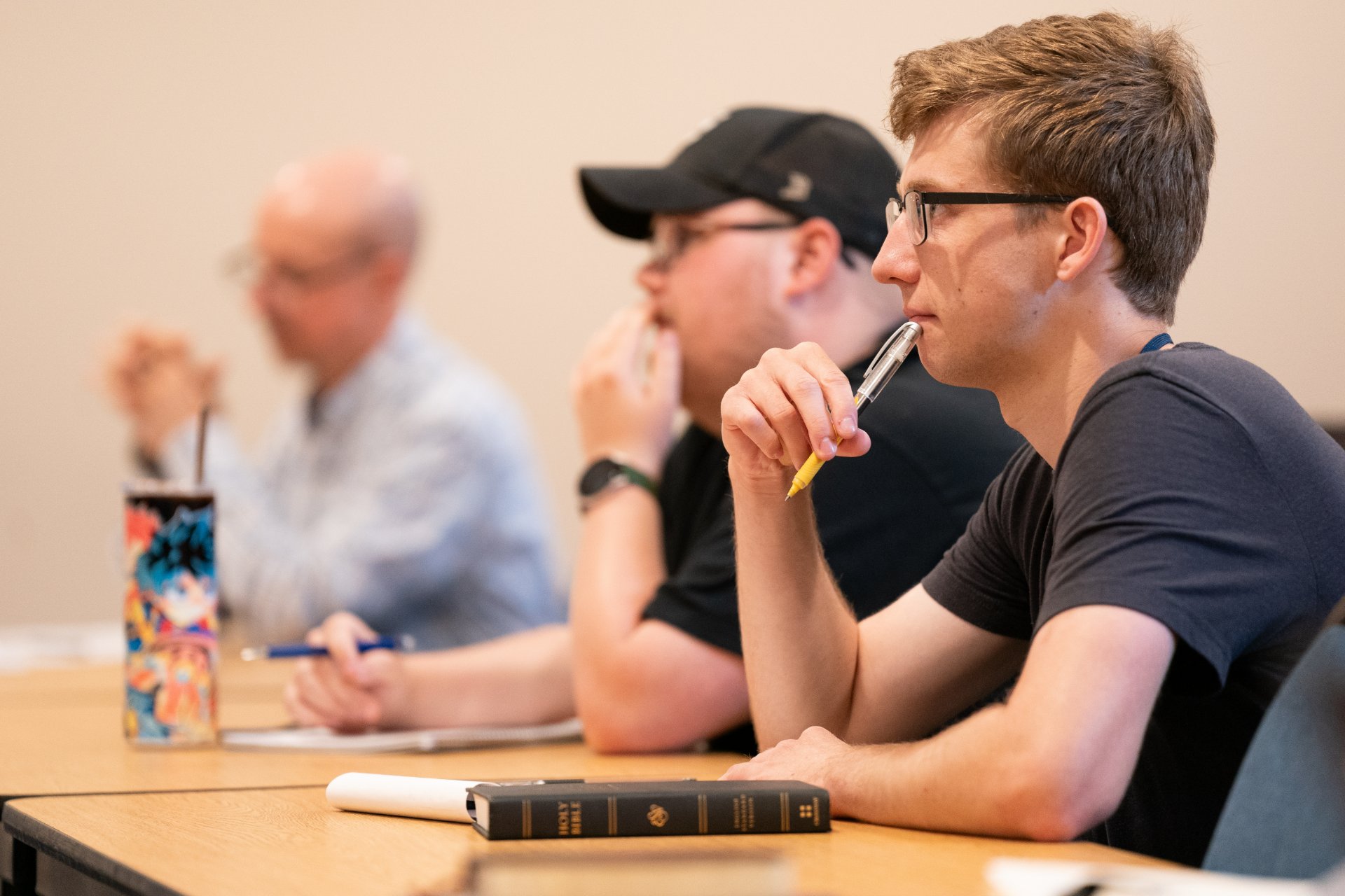 Three students sit in a row at a classroom table, pens in hand, focused on a lecture. A Holy Bible rests on the desk in front of the nearest student.