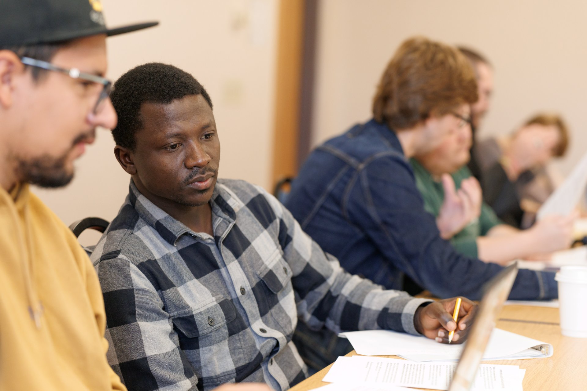 A close-up of a row of seminary students seated at a table during class. In the foreground, a student in a plaid shirt holds a pencil and looks attentively forward, while fellow students in the background participate in discussion.