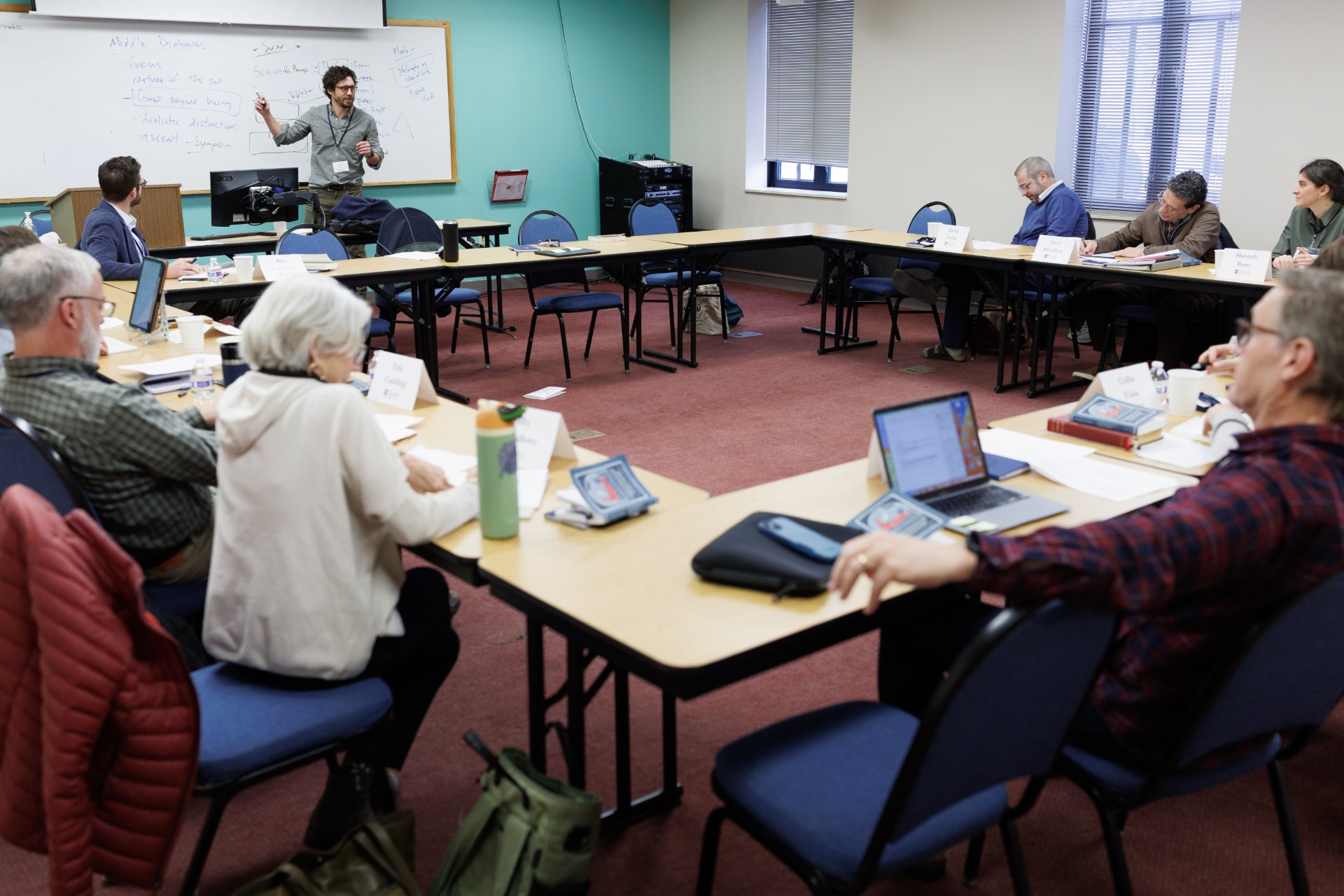 A professor writes on a whiteboard covered in course notes as roughly ten adult students follow along from laptops, books, and printed materials arranged around a U-shaped table configuration in a seminary classroom.
