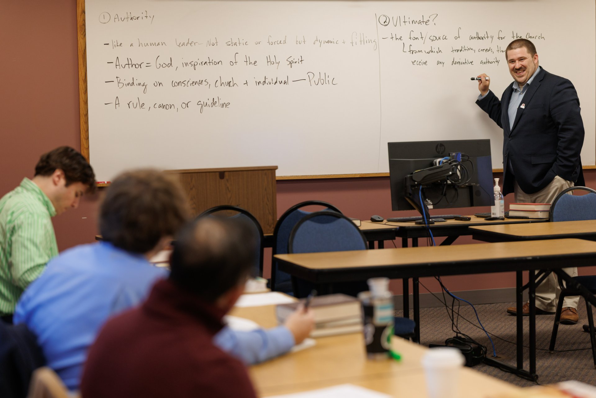 A smiling instructor writes on a whiteboard covering topics of scriptural authority—including notes on God as Author, Holy Spirit inspiration, and the church's ultimate source of authority—while several students seated at tables take notes.