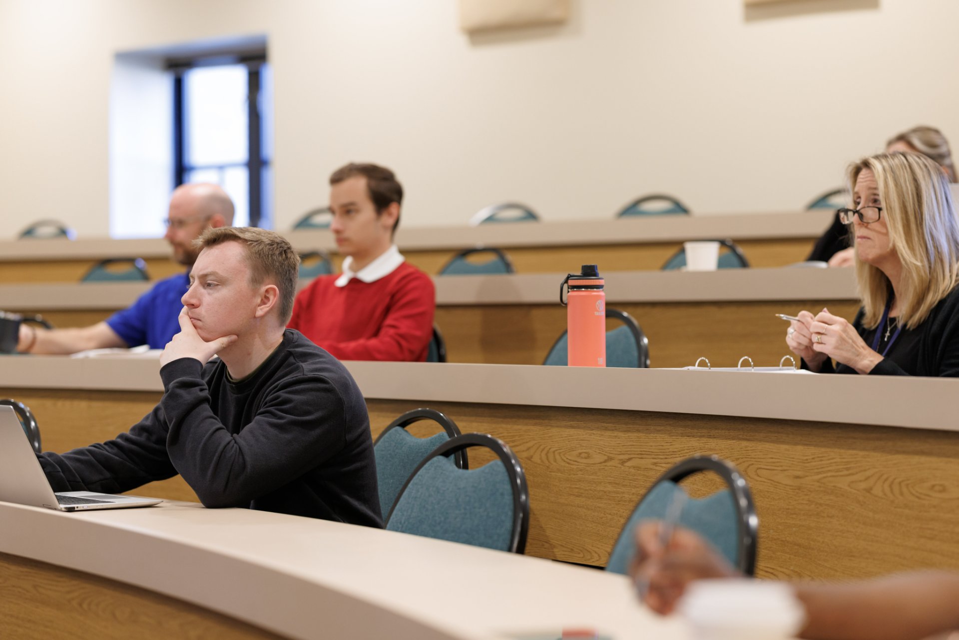 Four students of varying ages sit in a tiered lecture hall with wood-paneled desk rows, laptops and binders open, focused on content outside the frame.