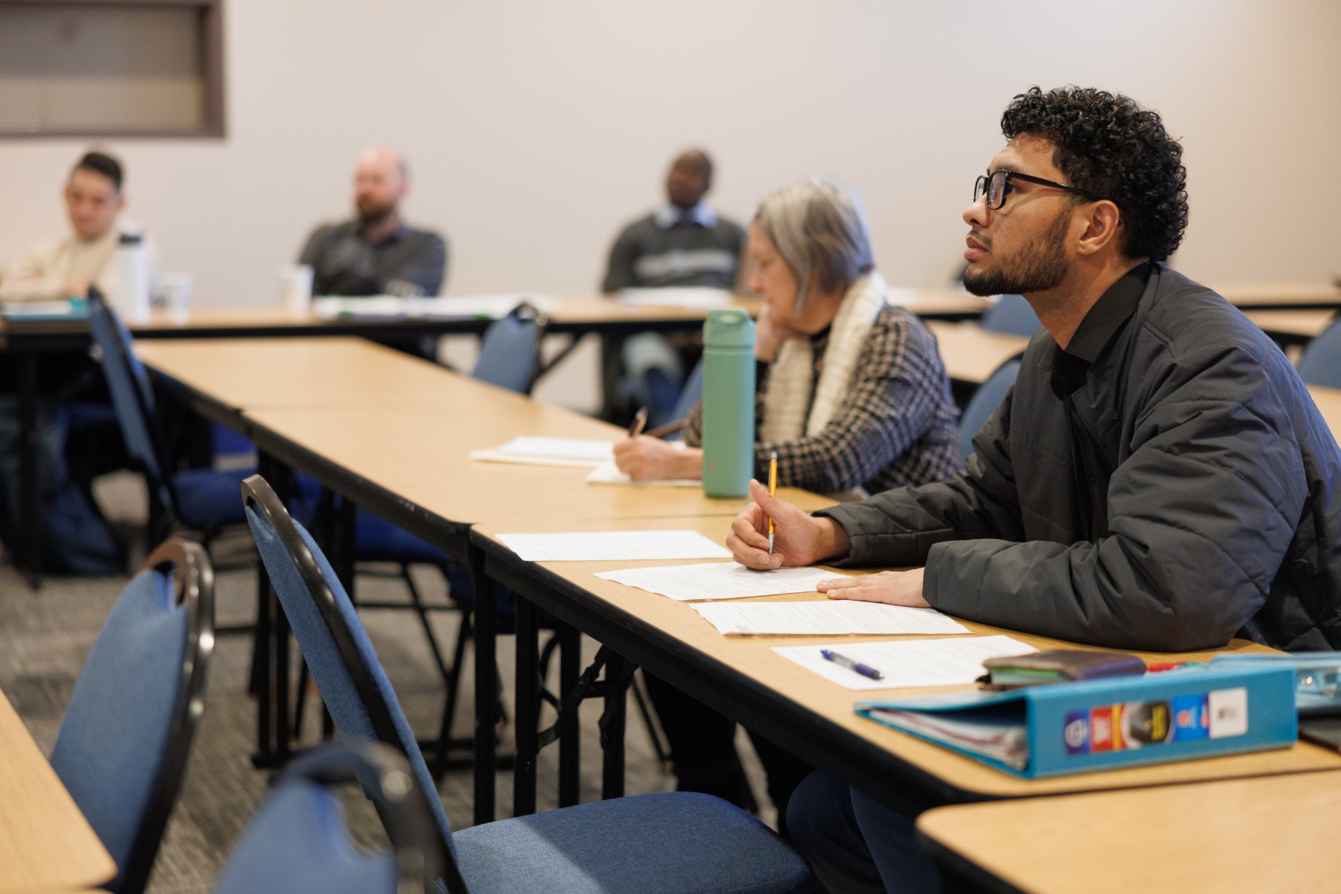 A young man wearing glasses and holding a pencil sits attentively at the front of a seminary classroom, with four other students of varying ages visible in the rows behind him.