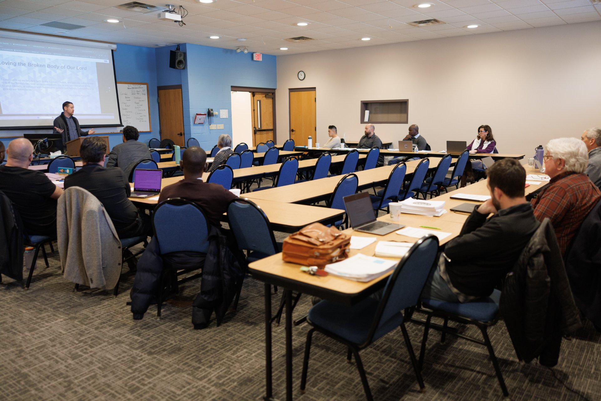 A diverse group of adult students with laptops and printed materials face an instructor at a podium; the projected slide reads 'Loving the Broken Body of Our Lord.'