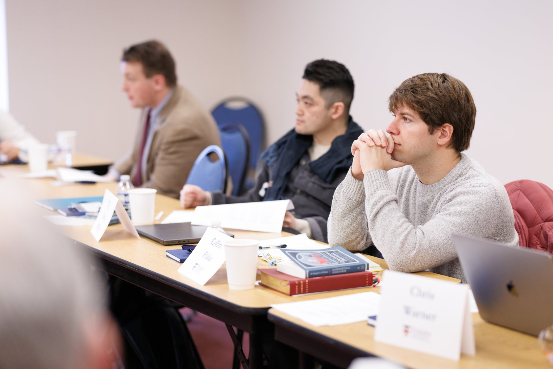 Three male students sit at a long classroom table with name placards, textbooks, laptops, and coffee cups, focused on an unseen instructor or speaker.