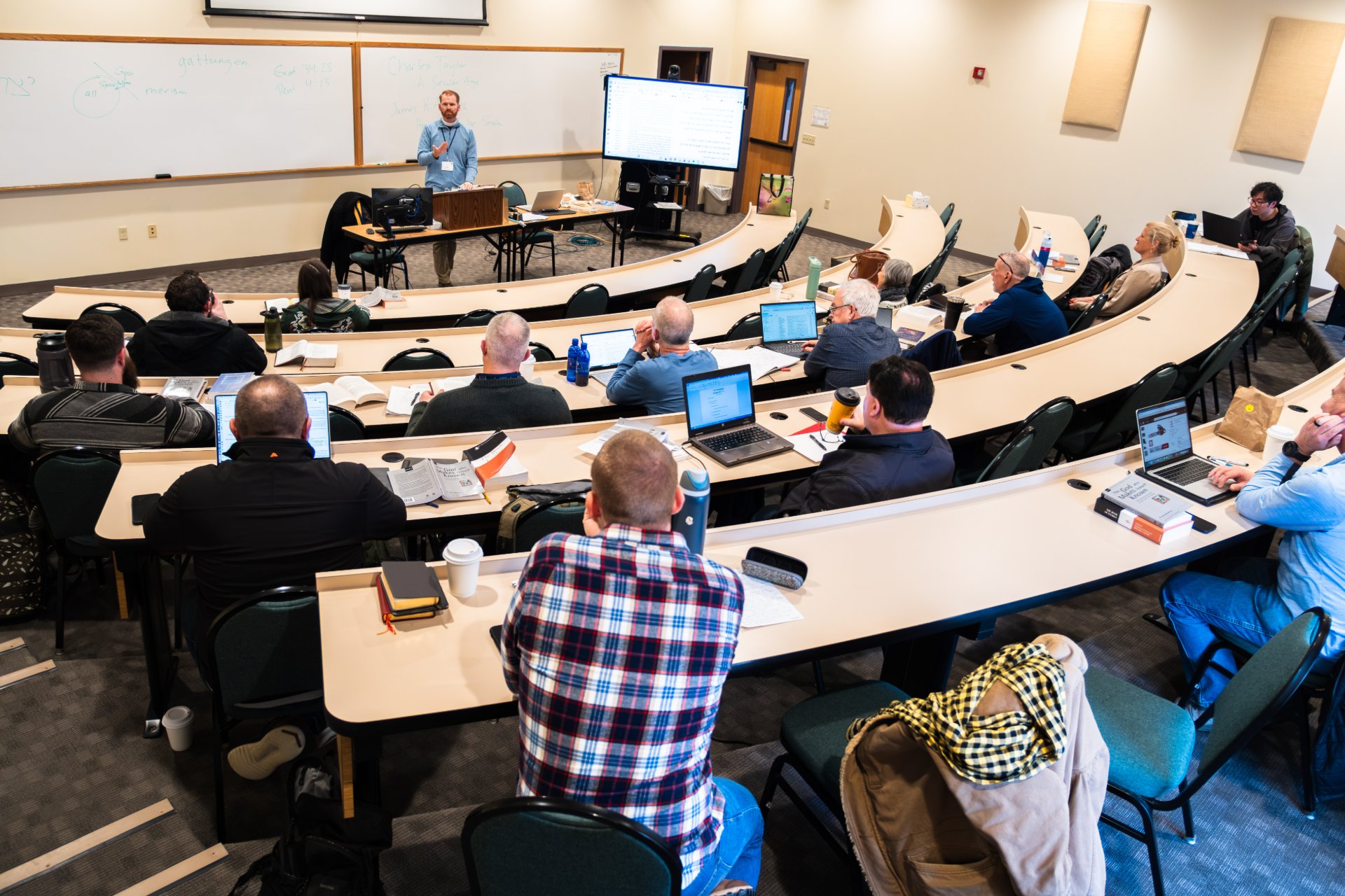 A professor stands at the front of a curved, tiered lecture hall, speaking before a whiteboard and projected screen. Roughly fifteen students face him with open Bibles, notebooks, and laptops on the desks before them.