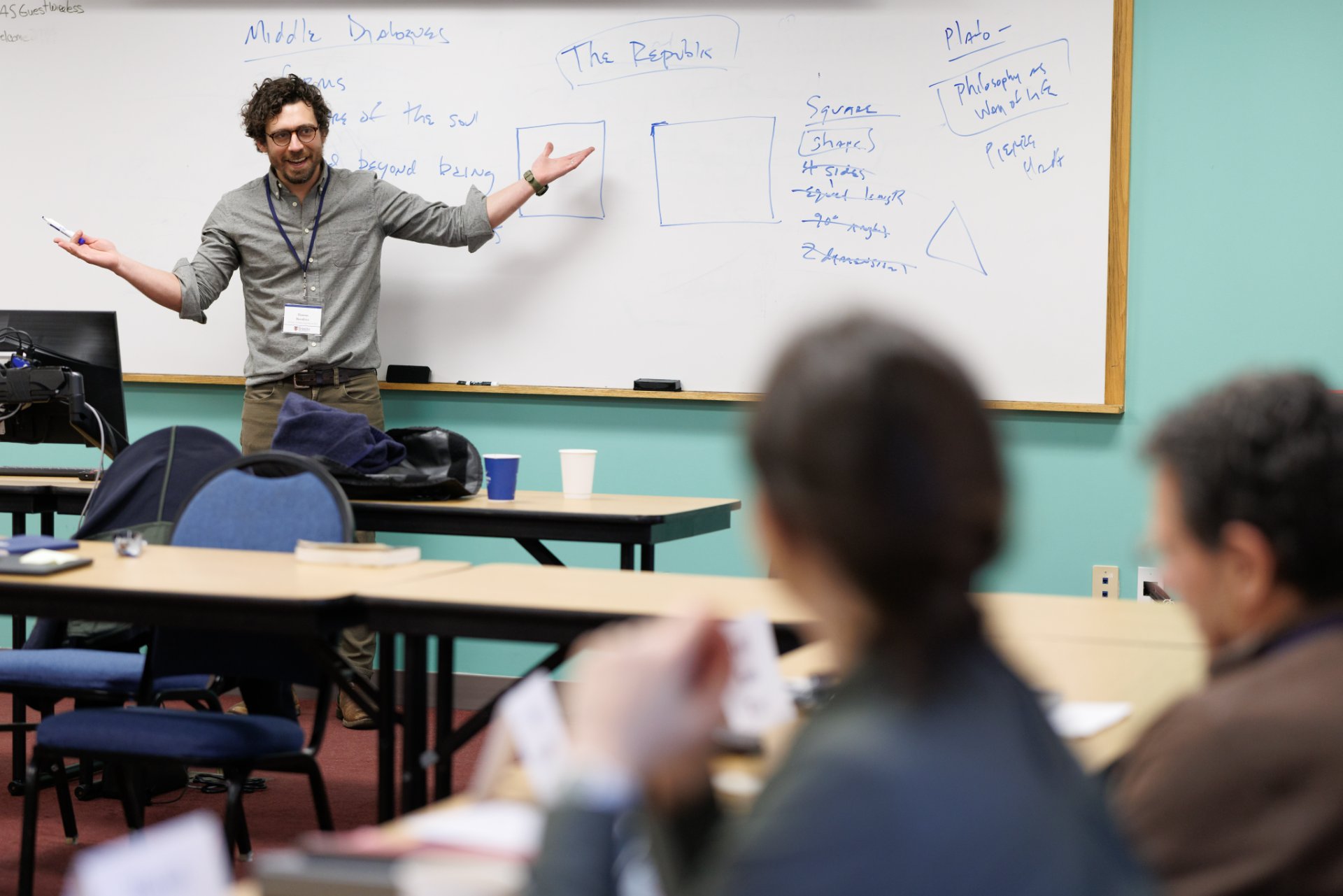 A male instructor wearing a name badge gestures expressively at a whiteboard covered with notes on Plato, including headings for 'The Republic,' 'Middle Dialogues,' and 'Philosophy as Way of Life,' while two students sit in the foreground with books and notes.