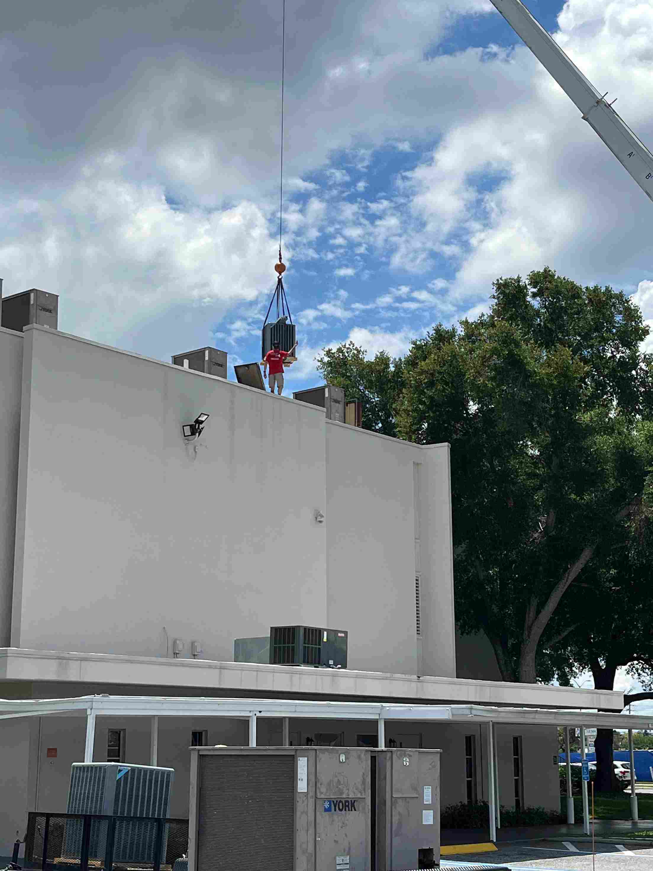 Crane lifting HVAC unit onto white building roof with trees