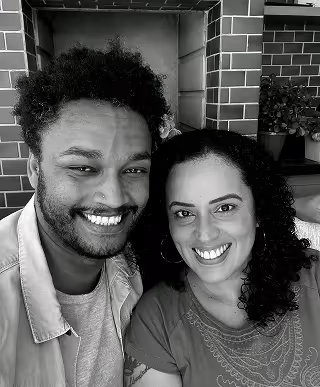 A smiling man and woman pose closely together for a selfie. The man has short curly hair, a beard, and a nose ring, while the woman has long curly hair and is wearing hoop earrings. They are sitting in front of a tiled wall with potted plants in the background. The image is black and white and cropped into a circular frame.