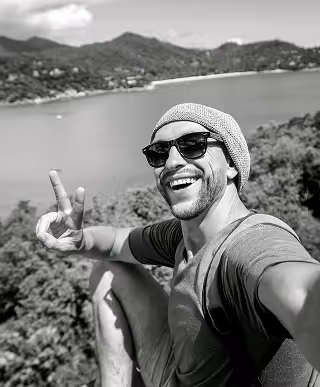 Man wearing sunglasses and a beanie making a peace sign while taking a selfie with mountains and water in the background.
