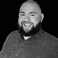 Smiling bald man with a beard wearing a patterned button-up shirt against a black background.