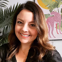 Smiling woman with wavy brown hair sitting indoors in front of plant and colorful artwork.