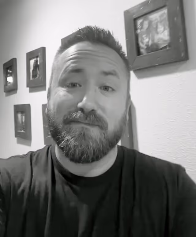 Black and white portrait of a bearded man with short hair wearing a dark t-shirt, standing in front of a wall with framed pictures.