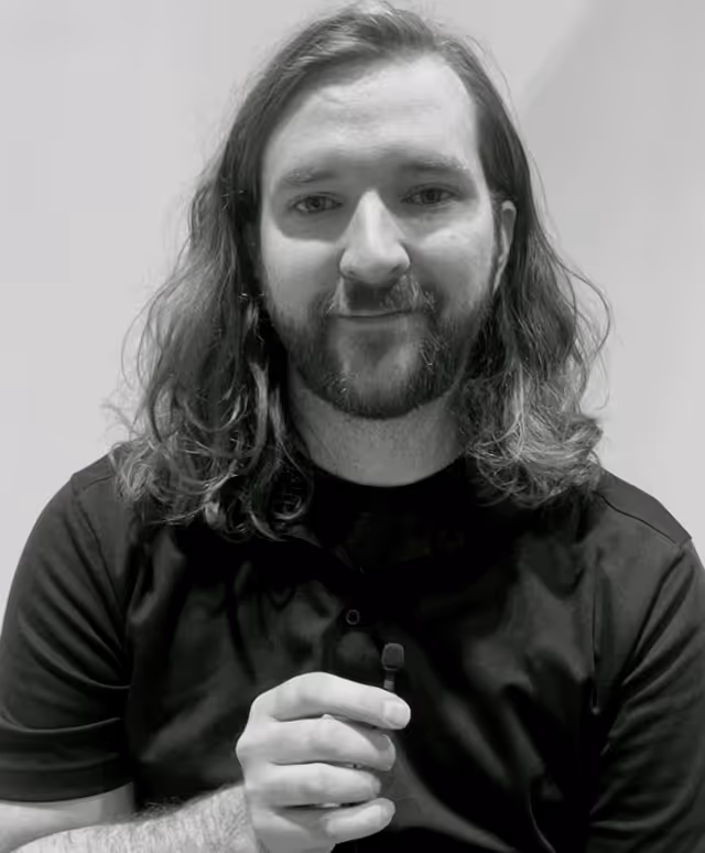 Man with shoulder-length hair and beard, wearing a black shirt and holding a microphone.