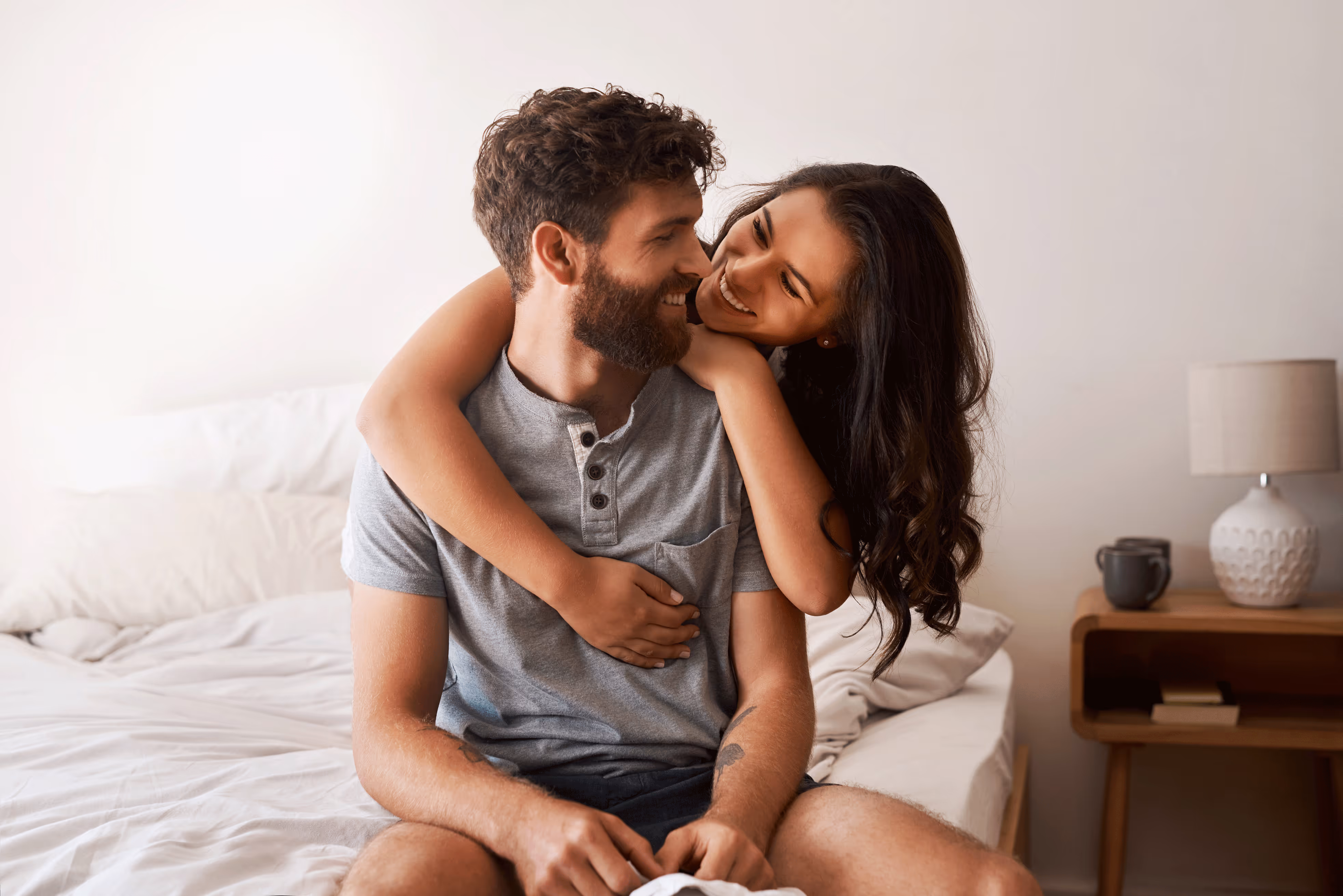 Smiling woman with arms around a seated man, both sharing a joyful moment on a bed in a bright bedroom.